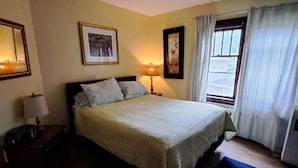 A hotel room with a bed, two bedside lamps, a window with curtains, and a framed picture on the wall. - Downtown heritage flat near everything (Dartmouth)