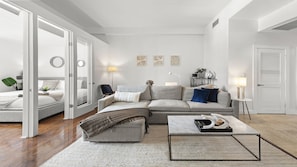 Uma sala de estar moderna com sofá, mesa de centro e mesa lateral. Há uma porta que dá para outro cômodo e uma parede com três objetos decorativos.