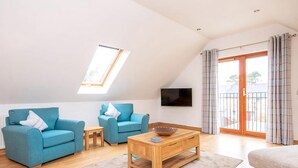 A modern living room with a sloped ceiling, two blue sofas, a wooden coffee table, and a TV mounted on the wall. - 2 bedroom accommodation in Dornoch (Dornoch)