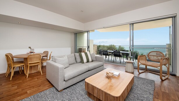 Una sala de estar moderna con un sofá gris, una mesa de centro de madera y sillas de mimbre. Hay una zona de comedor con una mesa y sillas de madera. La habitación tiene grandes puertas de cristal que dan a una terraza exterior con vistas al mar.
