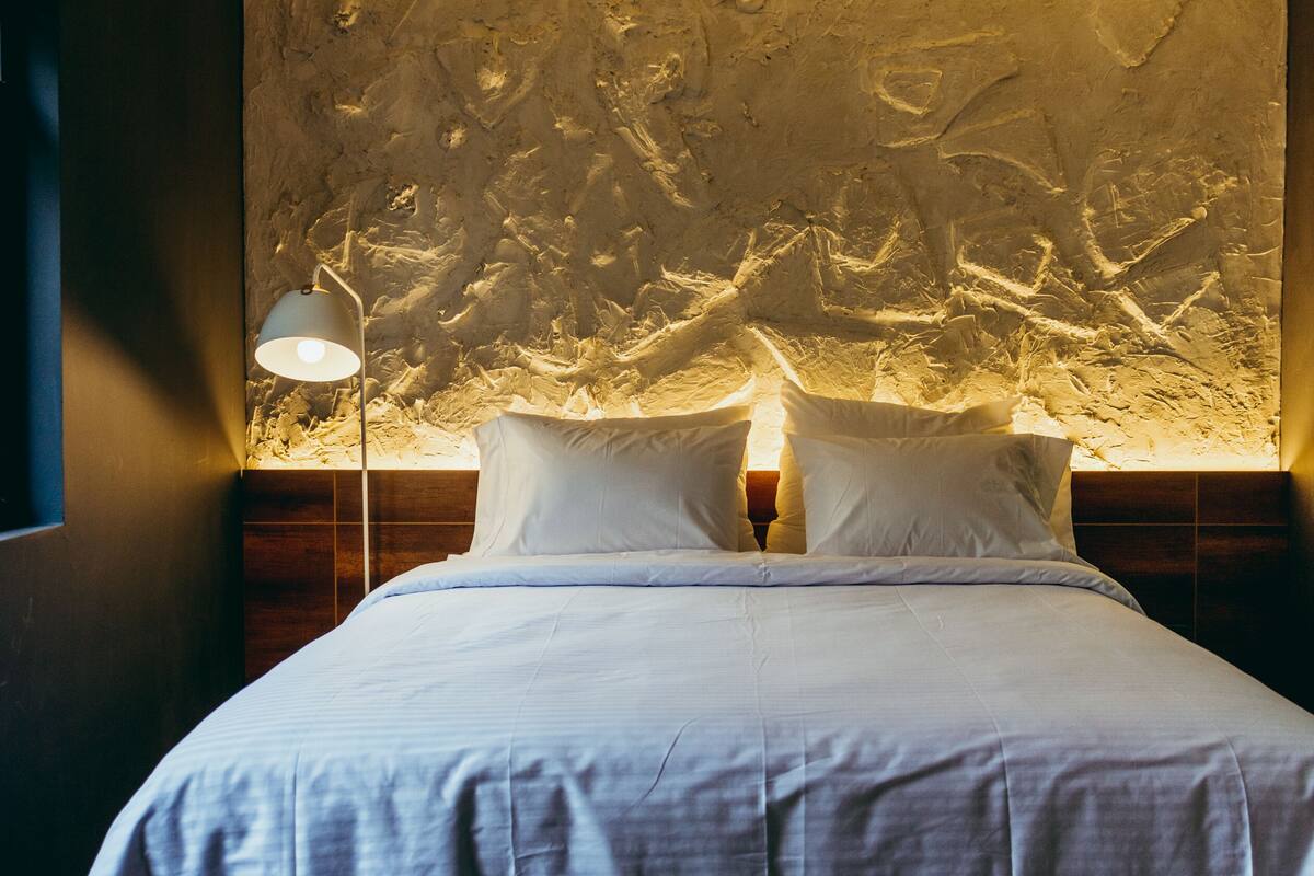 A bed with white linens in a room with a textured wall and a nightstand with a lamp.