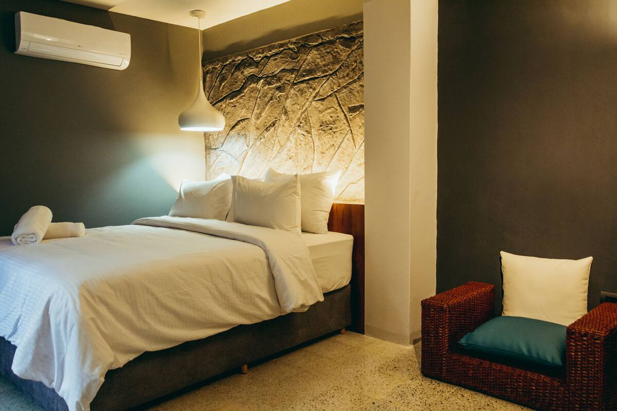 A bedroom with a bed, a nightstand, a wall-mounted air conditioner, a lamp, and a wicker chair.
