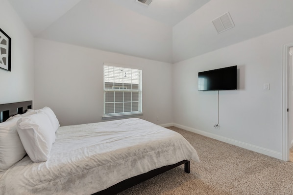 A bedroom with a bed, a window with blinds, a television mounted on the wall, and a door.