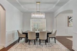 Une salle à manger avec un lustre, une grande fenêtre équipée de stores et un tableau accroché au mur.