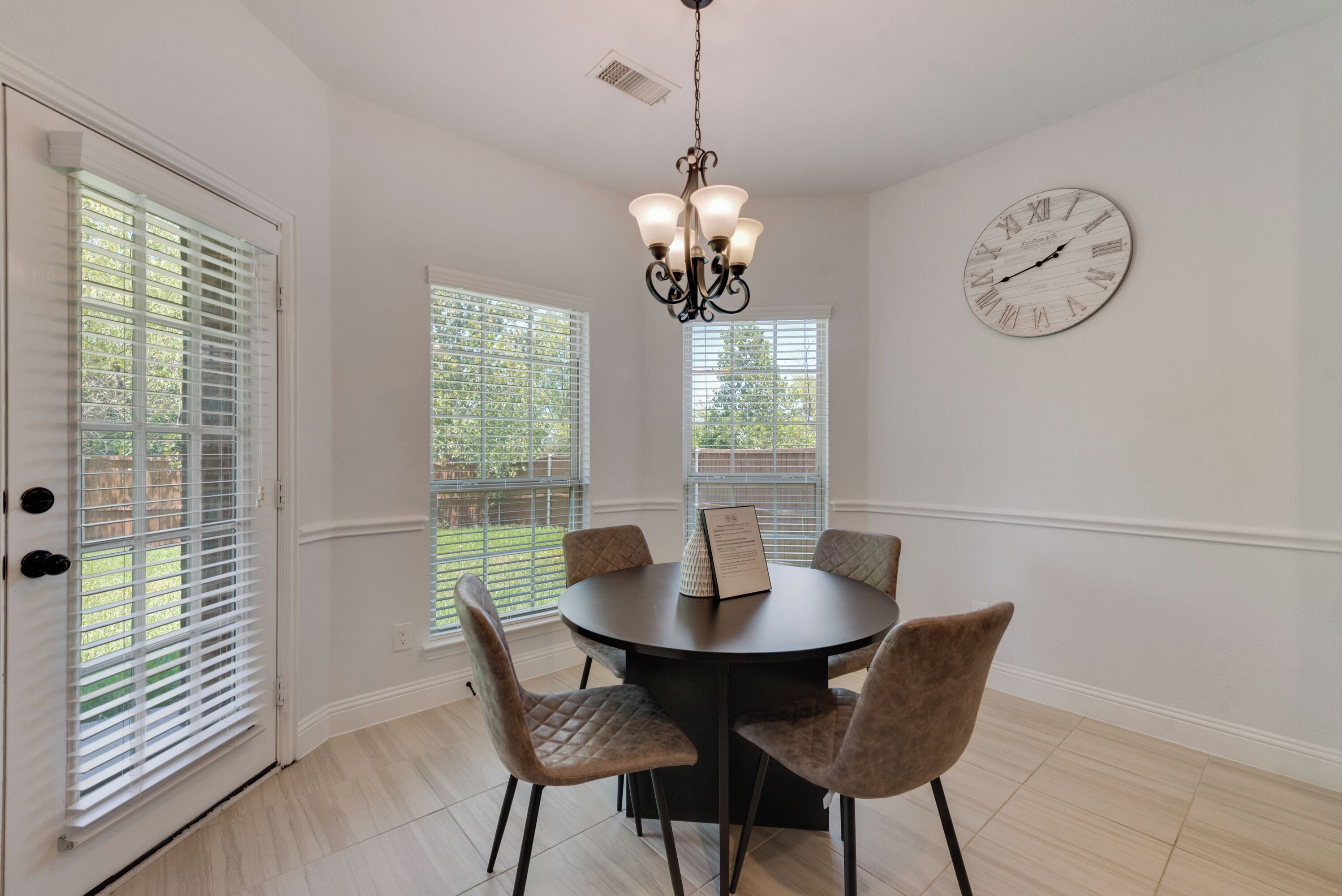 A dining area with a round table, four chairs, a chandelier, a wall clock, and large windows with blinds.