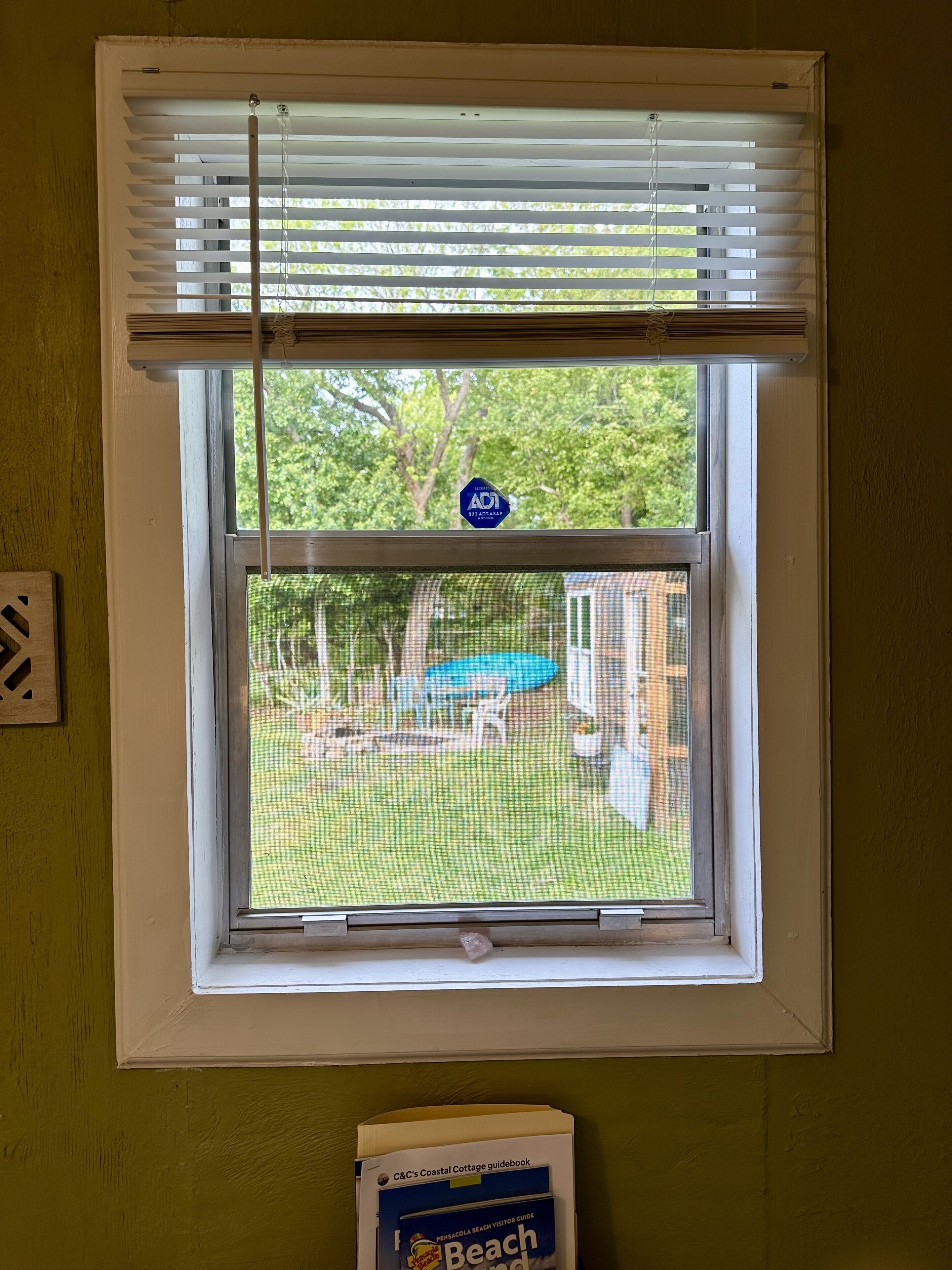 A window with a view of a backyard, a pool, and a beach guidebook on the windowsill.