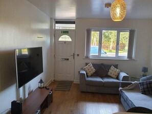 A living room with a flat-screen TV, a grey sofa with patterned cushions, a window with a view of trees, and a fire exit sign. - Immaculate 2-bed Apartment in Leigh (Leigh)