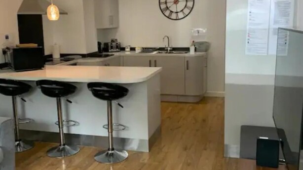 A modern kitchen with a central island, bar stools, pendant lighting, and a wall clock. - Immaculate 2-bed Apartment in Leigh (Leigh)