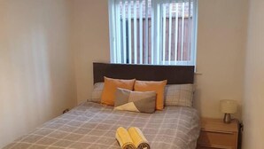 A neatly made bed with a checkered bedspread, two pillows, and rolled towels. There is a bedside table with a lamp and a window with blinds. - Immaculate 2-bed Apartment in Leigh (Leigh)