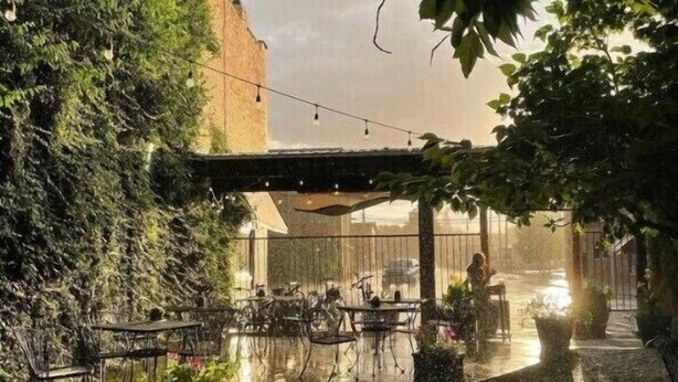 An outdoor café with tables and chairs under a pergola, surrounded by greenery and a brick wall.