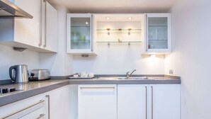 Une cuisine moderne avec des meubles blancs, une bouilloire en acier inoxydable, un grille-pain et un meuble vitré au-dessus de l’évier.
