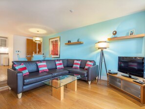 A modern living room with a sectional sofa, a glass coffee table, a TV, and a floor lamp. - 208 By the Bridge Apartment (Inverness)
