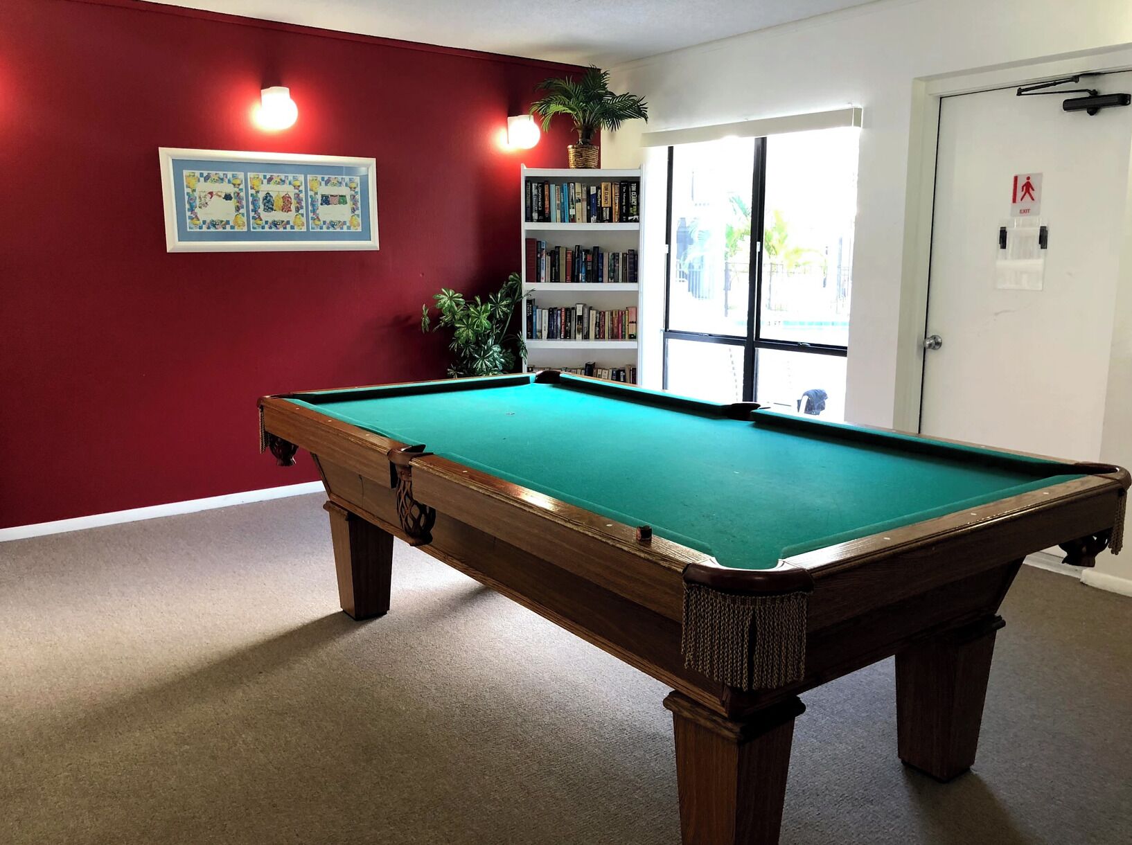 Une table de billard dans une pièce aux murs rouges, avec une bibliothèque et une porte sur laquelle est apposée un panneau indiquant la sortie.