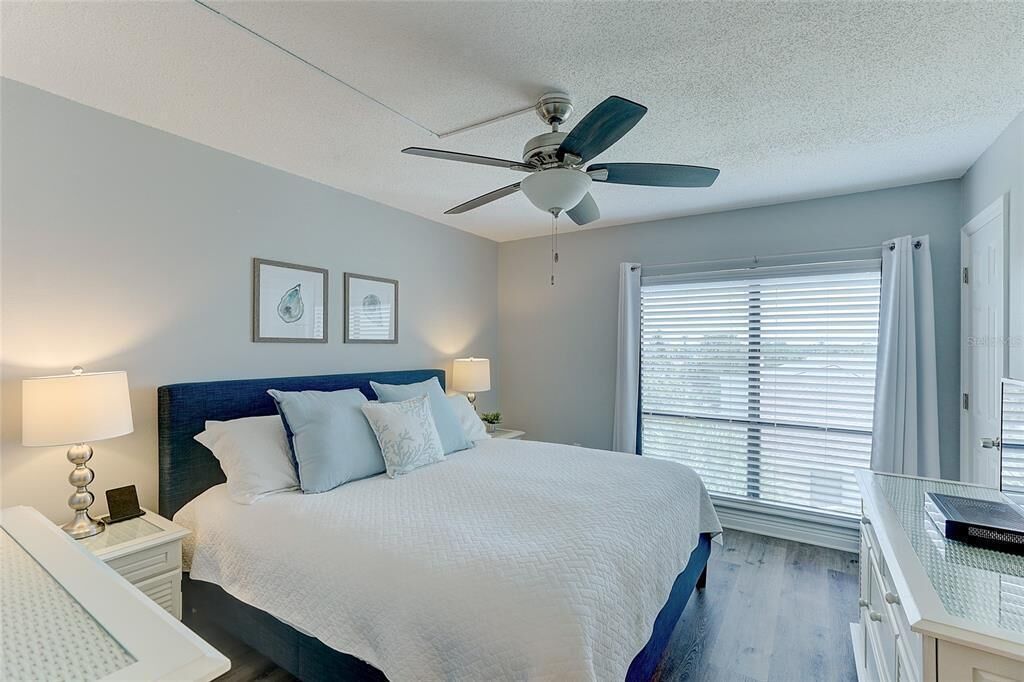 A bedroom with a bed, two bedside lamps, a ceiling fan, a window with blinds, and a desk with a laptop.