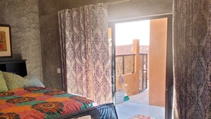 A room with a bed, a sofa, and a table with a remote control. There is a window with curtains and a view of the outside.