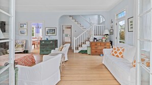 A spacious living room with a staircase leading to another level, white furniture, and a wooden floor. - Charming Coastal Biddeford Pool Home (Biddeford)