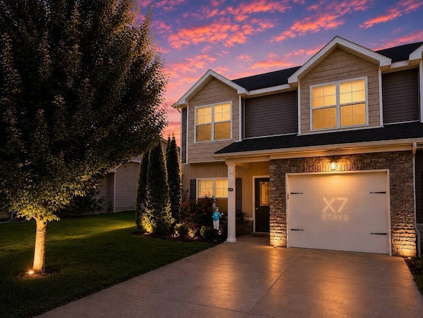 A two-story house with a garage, a well-lit driveway, and a tree in the front yard.
