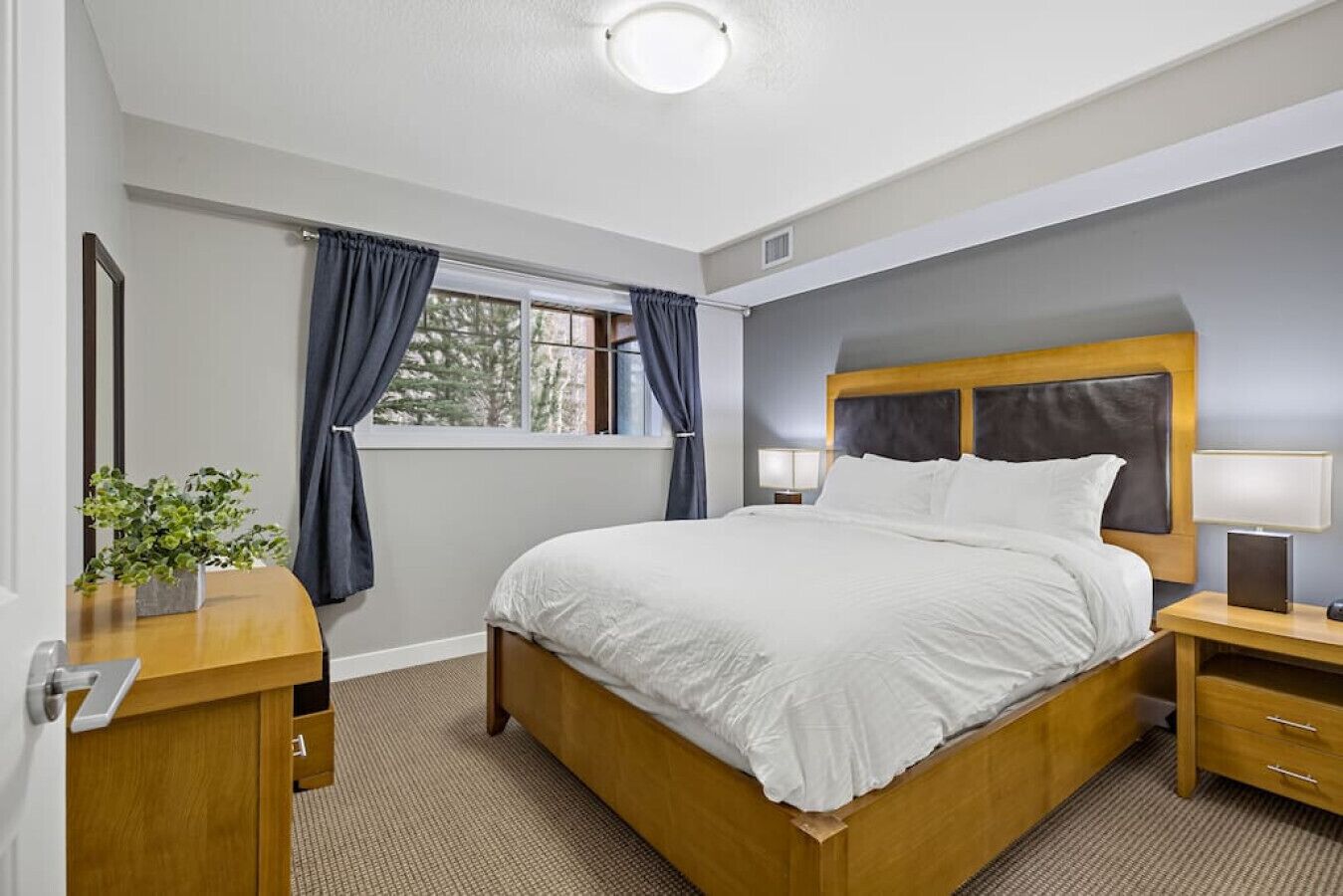 Ein ordentlich eingerichtetes Schlafzimmer mit einem großen Bett, Nachttischen, einem Fenster mit Vorhängen und einer kleinen Pflanze auf dem Nachttisch.