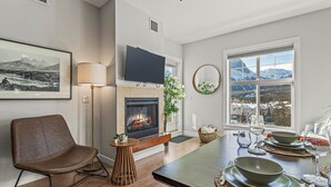 A modern living room with a fireplace, a flat-screen TV, a dining table set for two, and a view of mountains through a large window.