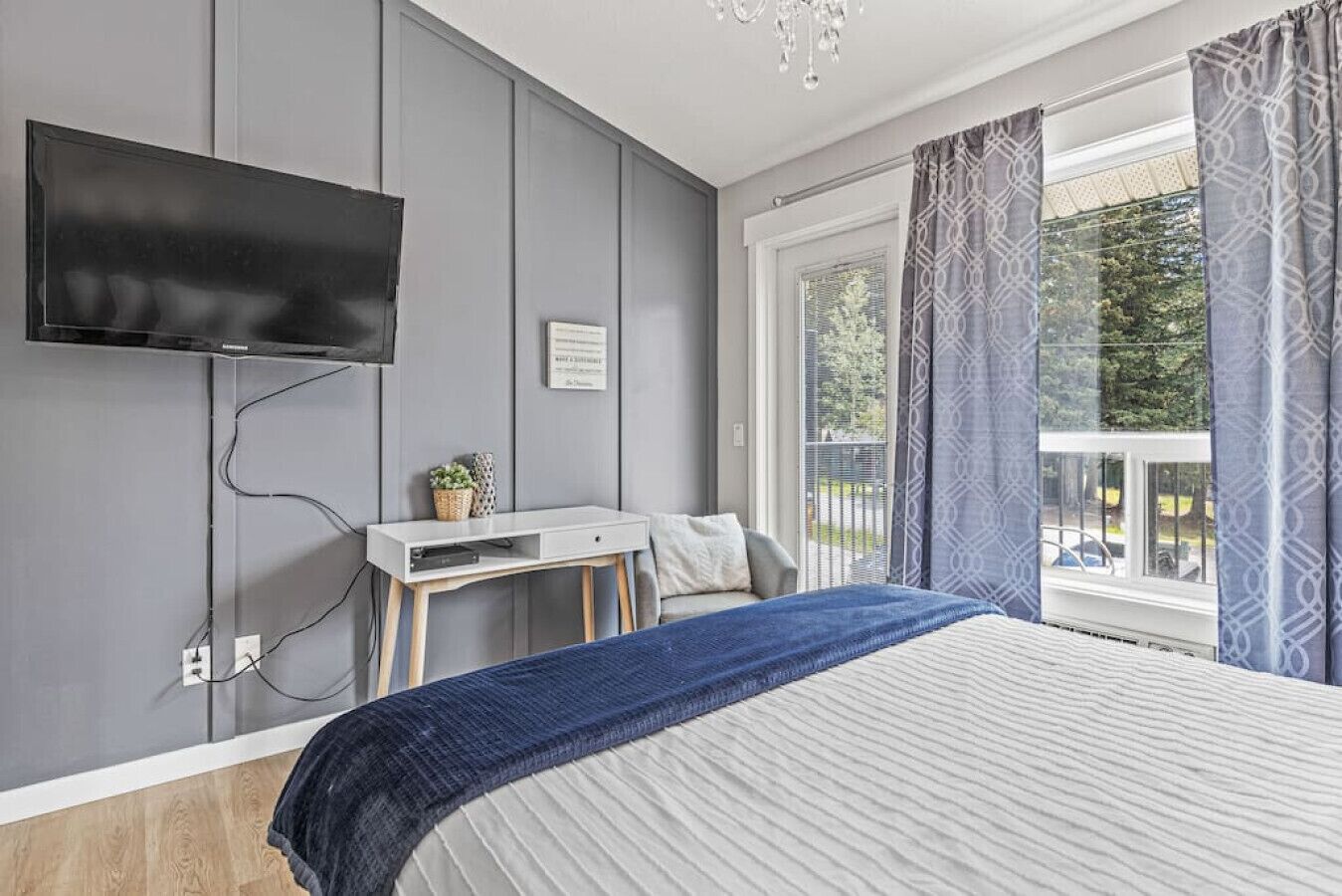 A bedroom with a bed, a desk, a TV mounted on the wall, and a window with curtains.