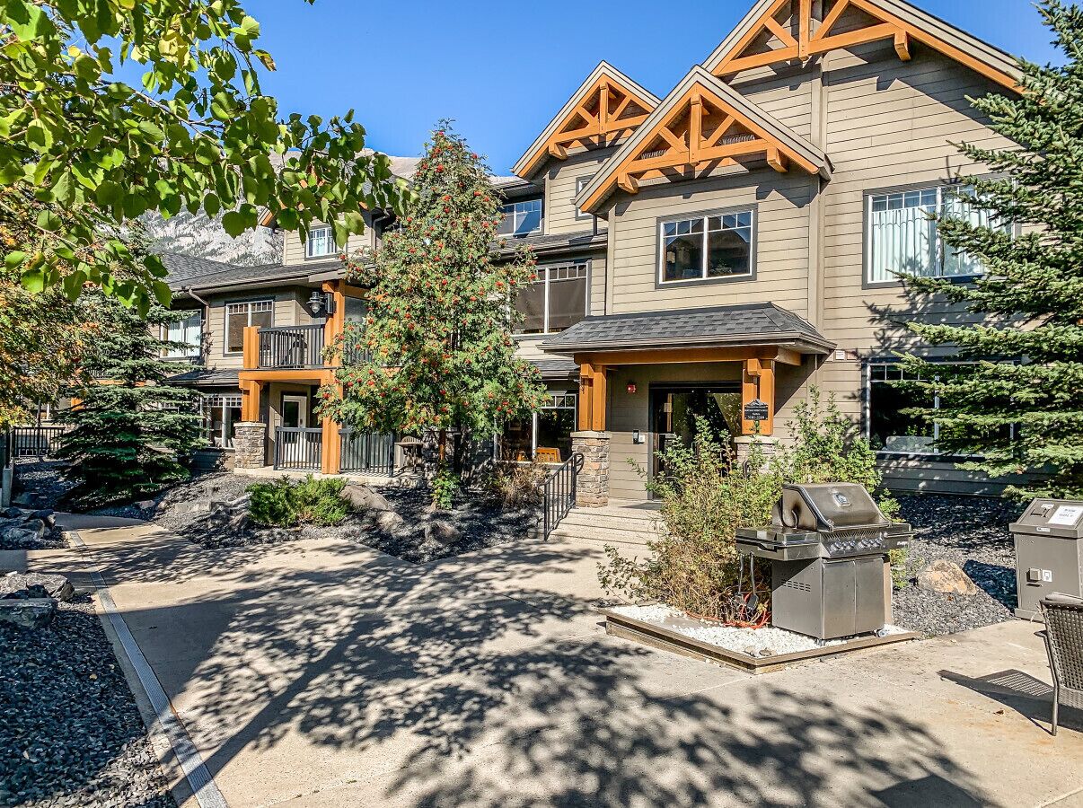 A two-story house with a stone foundation, a wooden deck, and a barbecue grill in the front yard.