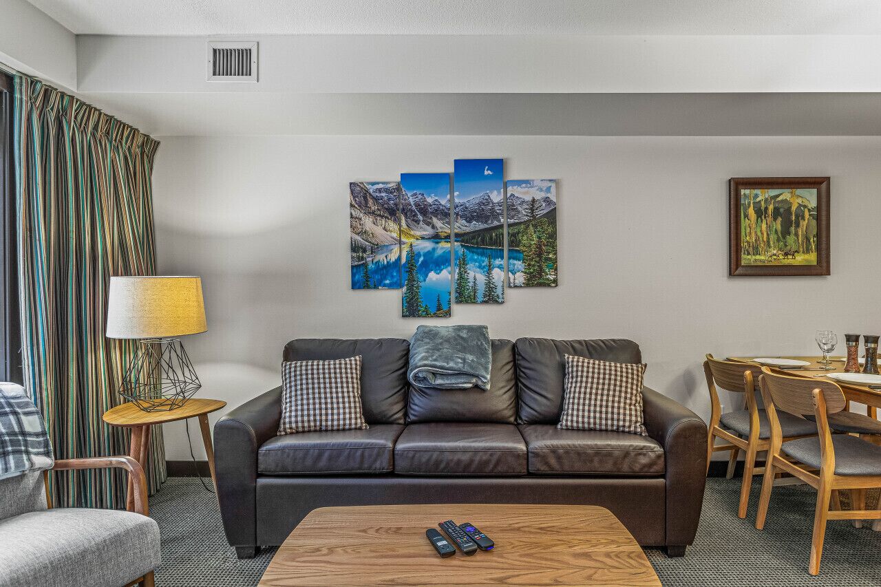 A modern living room with a leather sofa, a coffee table, a dining area with wooden chairs, and a large landscape painting on the wall.