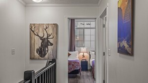A hallway with a wooden deer head artwork, a bed with a purple blanket, and a poster on the wall. - 2 Bed |Sleeps 8|Walkout to Heated Pool|Hot Tub (Canmore)