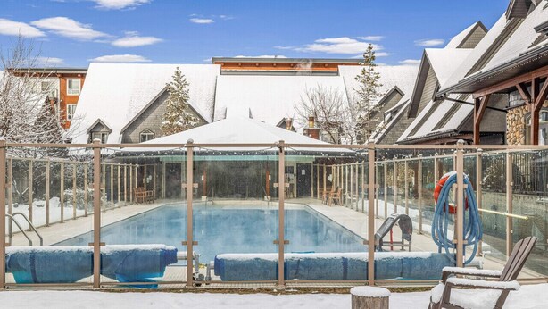A covered outdoor swimming pool with lounge chairs and a hose reel, surrounded by snow-covered trees and buildings. - 2 Bed |Sleeps 8|Walkout to Heated Pool|Hot Tub (Canmore)
