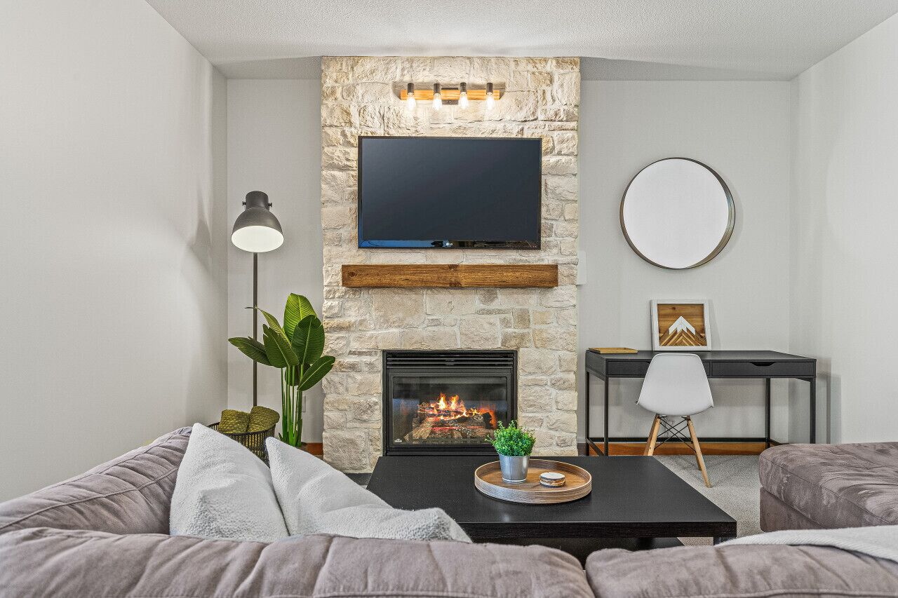 Ein Wohnzimmer mit Kamin aus Stein, einem darüber angebrachten Flachbildfernseher, einem runden Spiegel an der Wand und einem modernen Sofa.
