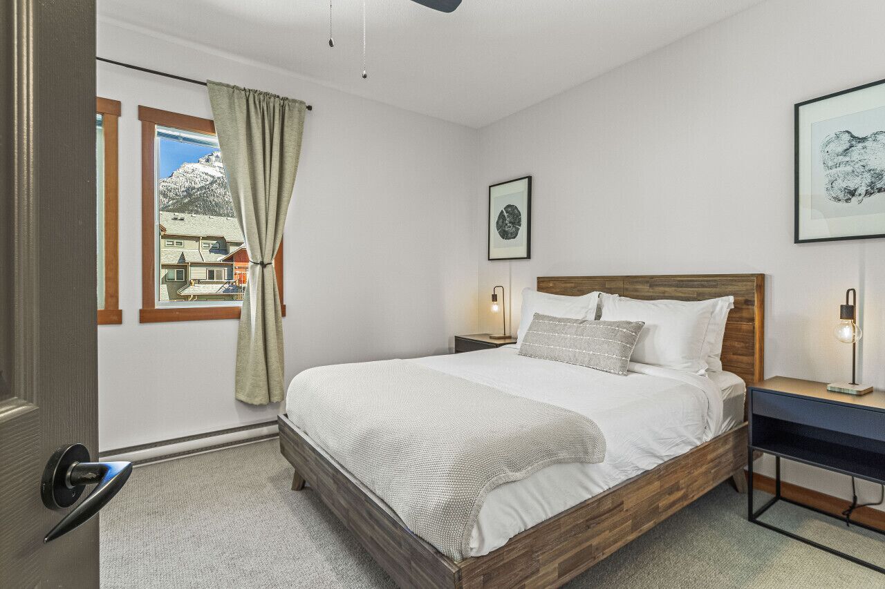 A bedroom with a wooden bed, a nightstand with a lamp, a window with a view of mountains, and a door with a handle.