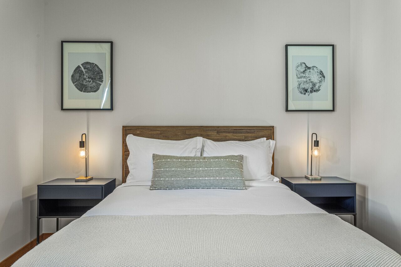 A neatly made bed with a white comforter and a patterned pillow, flanked by two bedside tables with lamps, each displaying a framed artwork on the wall above.