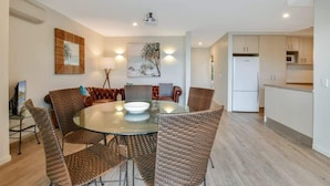 Una cocina moderna con armarios blancos, un refrigerador blanco y una zona de comedor con sillas de mimbre y una mesa de vidrio.
