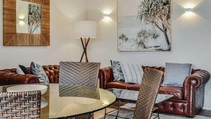 Una sala de estar con un sofá de cuero marrón, una mesa de centro de vidrio y una silla de mimbre. Hay una lámpara de pared y un cuadro que muestra una escena tropical.