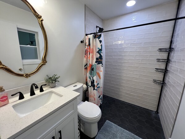 A bathroom with a white sink, a toilet, a shower with a floral curtain, a mirror with a gold frame, and a black towel rack. - Modern & Convenient 3/2 Home (Lake City)