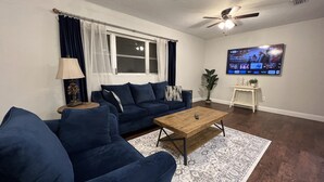 A living room with a dark blue sofa, a wooden coffee table, a TV mounted on the wall, and a ceiling fan. - Modern & Convenient 3/2 Home (Lake City)