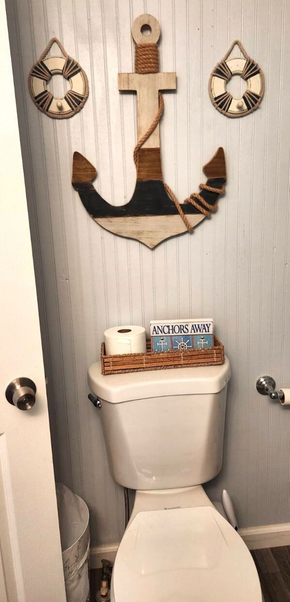 Des toilettes décorées d’une ancre en bois au-dessus et sur lesquelles est posé un panier portant l’inscription « ANCRES, HOISSEZ ! ».