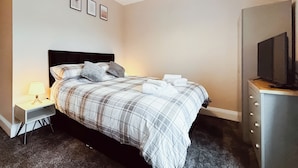 A bedroom with a bed, a nightstand, a lamp, a dresser, a television, and framed pictures on the wall.