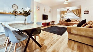 A modern living room with a dining area, a sofa, a TV, and a wall-mounted shelf with decorative items.