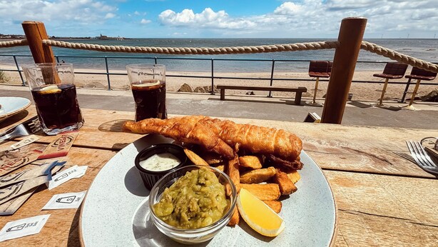 A meal of fish and chips with a drink and sauce on a wooden table by the beach.