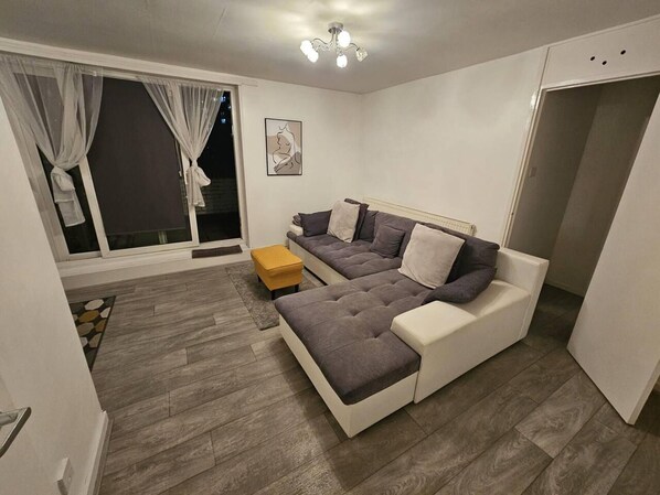 A modern living room with a grey sofa, a yellow ottoman, and a large window with curtains. - Stunning 1-bed Apartment in London (London)