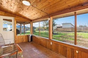 Un patio techado con paredes de madera y ventanas amplias, un bote de basura y una silla de metal.