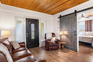 A room with a wooden ceiling, a brown leather couch, a dark wooden door, a sliding barn-style door, and a bed with a view. - Place on Pecan | Close to Main in Cowboy Capital (Bandera)