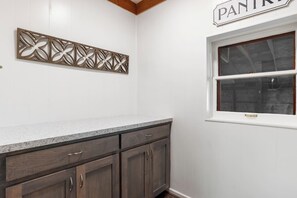 Een keuken met een granieten aanrecht, houten kastjes, een decoratief wandobject en een raam met een bordje 'PANTRY'.