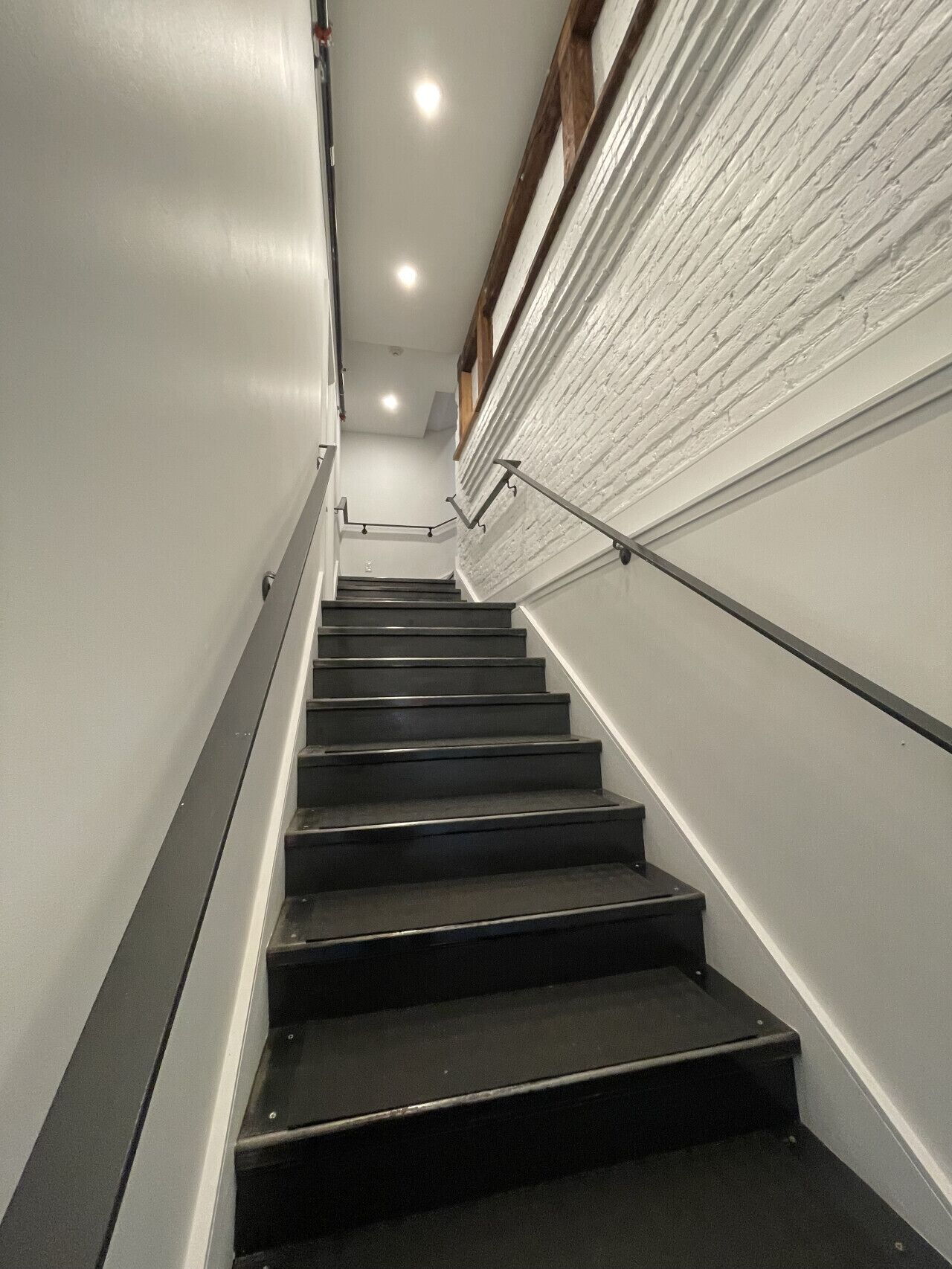 Eine Treppe mit schwarzen Stufen und Metallgeländern, die zu einem Zwischenpodest mit einer weißen Backsteinmauer und einem Holzbalken darüber führt.