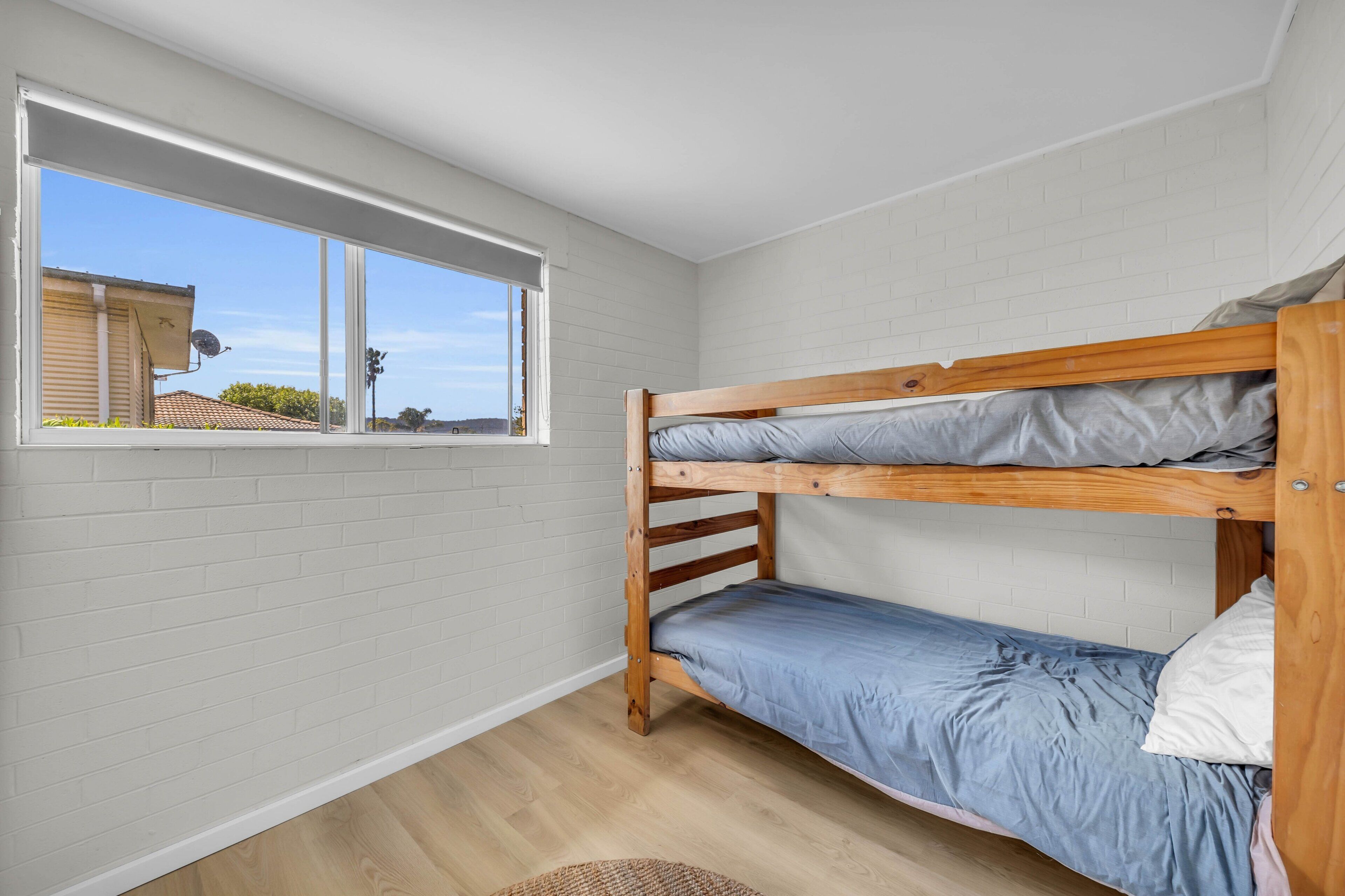 A room with a bunk bed, a window with a view of a house, and a white brick wall.