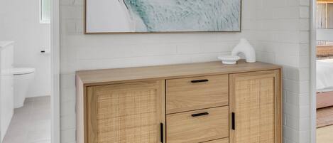 Un mueble de madera con cajones, un cuadro abstracto colgado en la pared y una pared de ladrillo blanco.