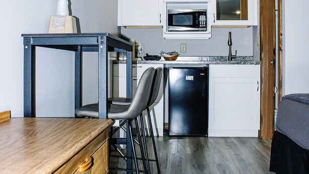 A compact kitchen with white cabinets, a microwave, and a dishwasher. There's a wooden table and a chair, and the floor is covered with grey wood-like flooring. - Renovated King Kitchenette (Branson)