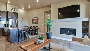 A modern living room with a fireplace, a TV mounted above it, and a dining area with a kitchen in the background.