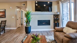 A living room with a fireplace, a television mounted above it, and a large window on the right. - Sage Creek Desert Escape C2 - Luxury Pool & HotTub (Moab)