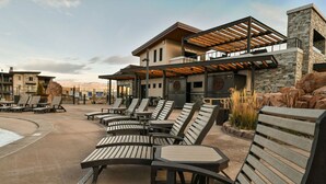 A modern outdoor seating area with lounge chairs and tables, a covered patio, and a stone building with a wooden deck. - Moab Arches C4 "Sage Creek" - Heated Pool-Hot Tubs (Moab)